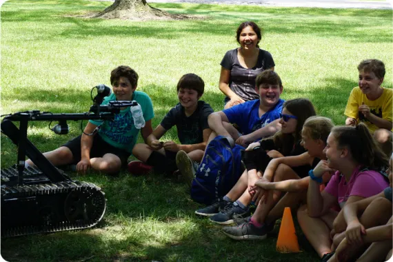 A group of kids sitting in the grass smiling and laughing as a small robot picks up a waterbottle